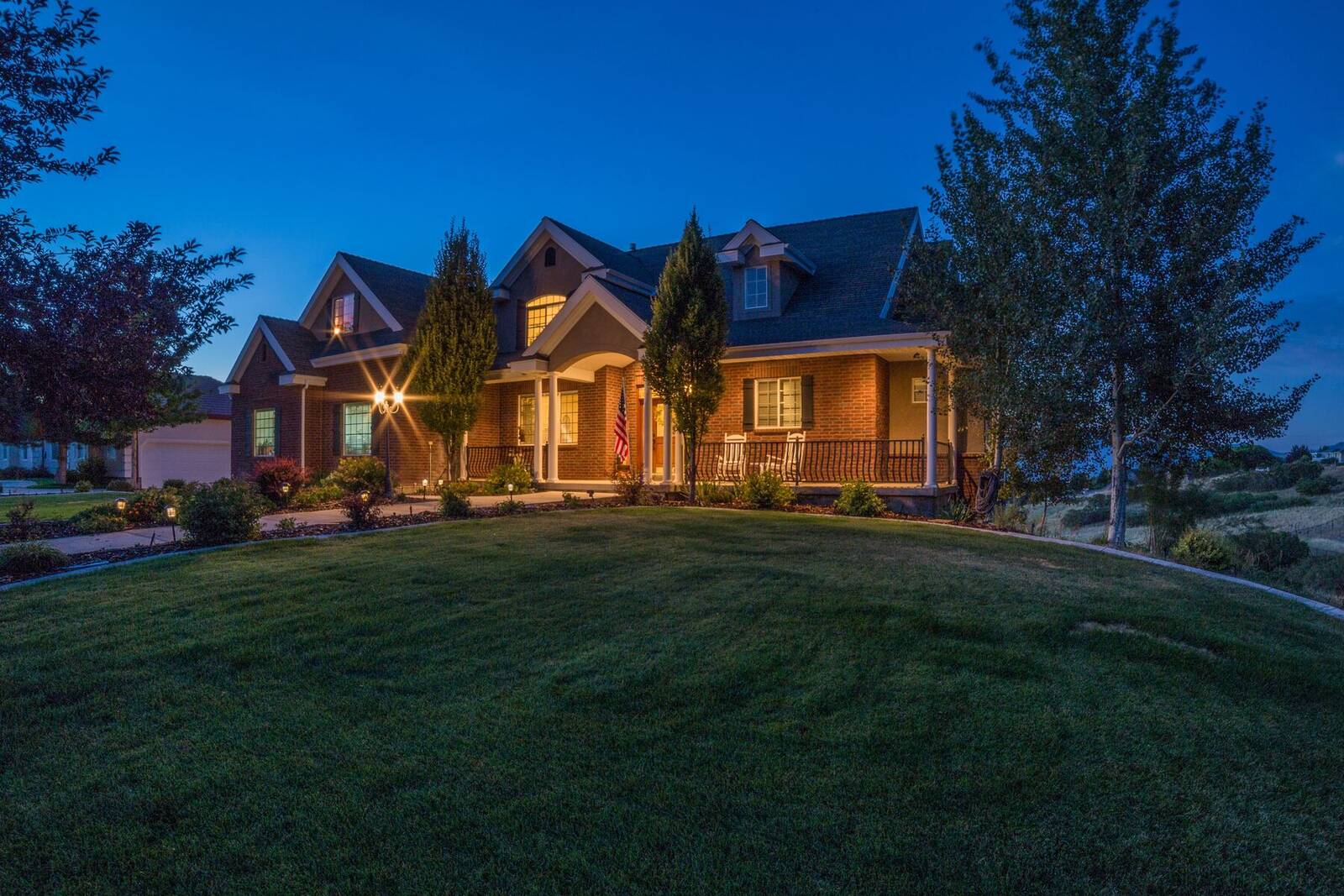A brick home with a wide front lawn and porch lights on at dusk