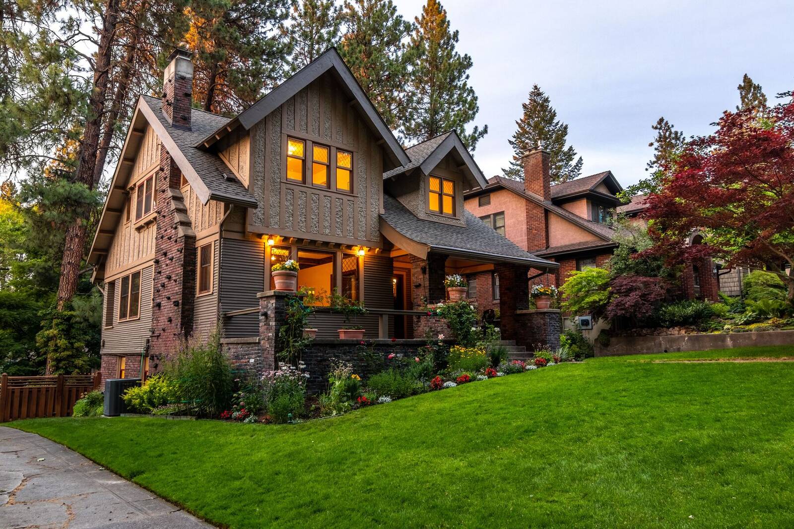 A two-story craftsman home with porch lights on, a manicured lawn and flower beds