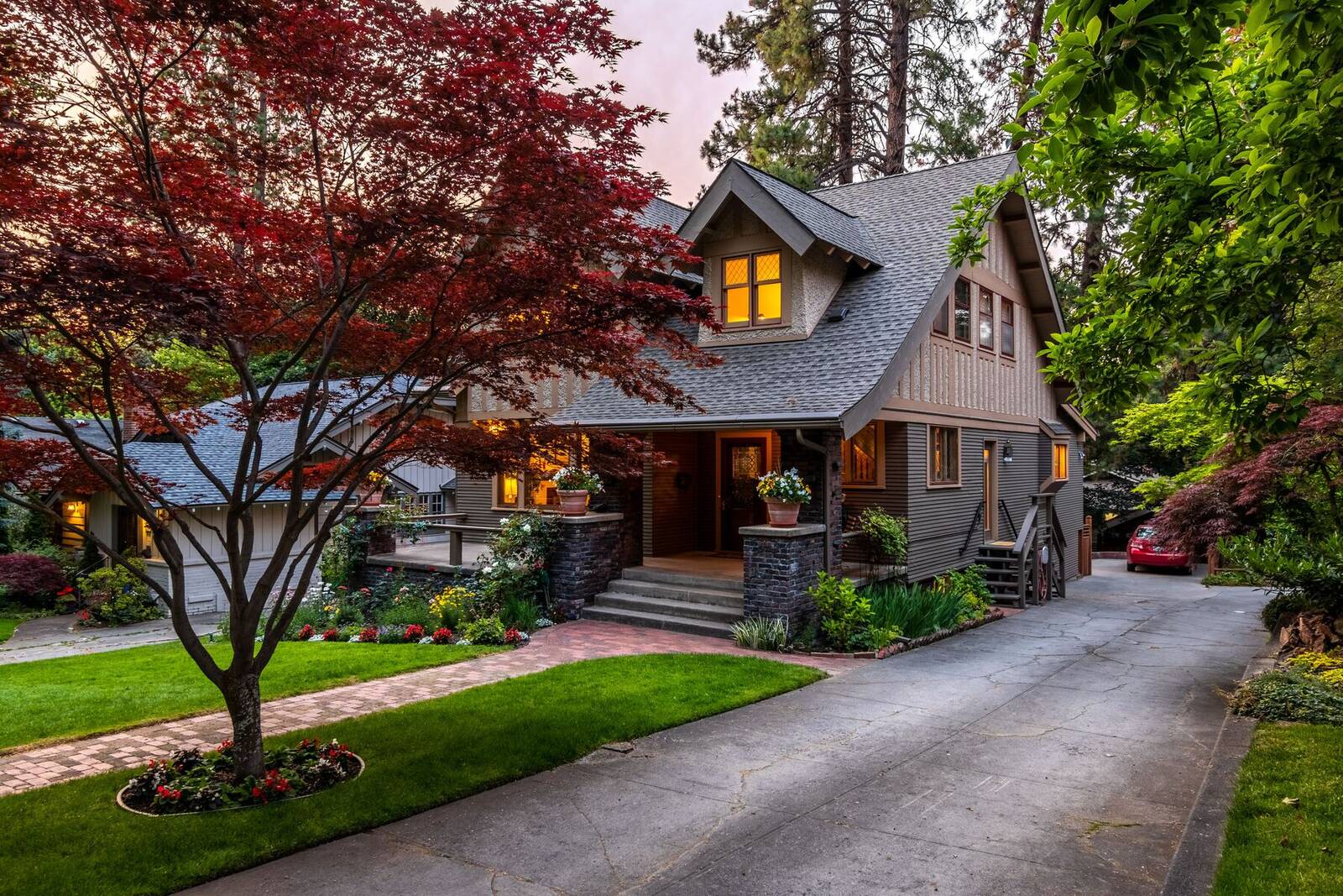 A craftsman-style home with a red Japanese maple, flower beds and lit windows at dusk